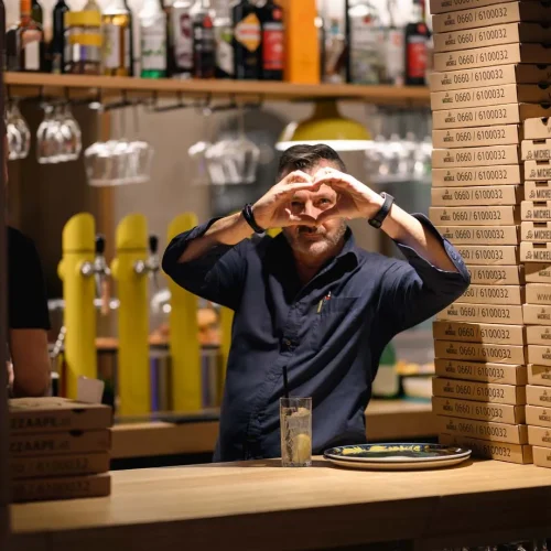 Am Bild zu sehen ist ein Kellner in der Pizzeria De Michele, der mit seinen Händen ein Herz formt und dabei in die Kamera blickt.